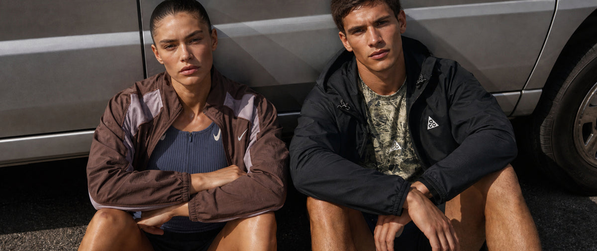 Two models sitting outdoors next to a silver car, wearing athletic jackets. The model on the left wears a brown Nike jacket over a blue top, and the model on the right wears a black Adidas jacket over a camouflage shirt.
