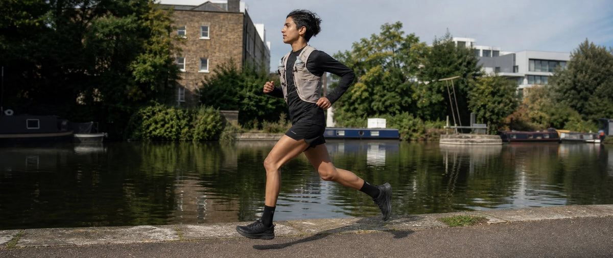 A woman running along a canal path wearing a gray and black athletic vest, black shorts, and black running shoes, with trees, water, and buildings in the background.