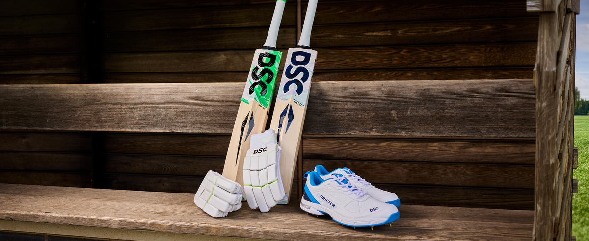 DSC cricket gear on a bench. a green and white bat, a white bat with white gloves and white and blue shoes Infront 