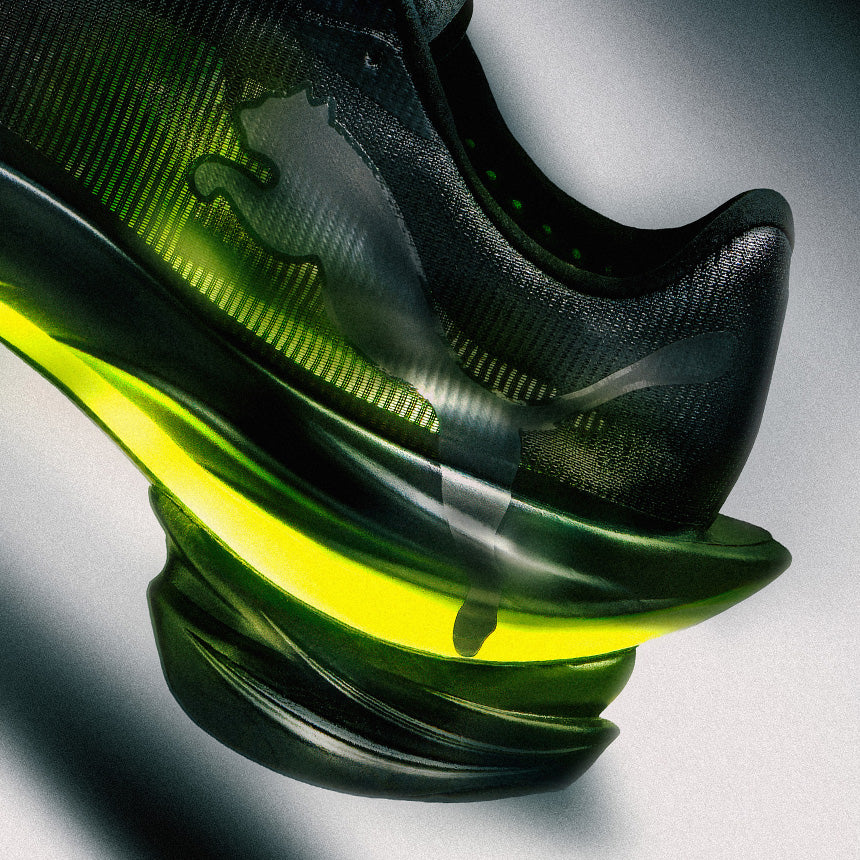 A close-up studio shot of a black athletic shoe with a translucent green and yellow sole, showing the heel and midsole details against a gray background.